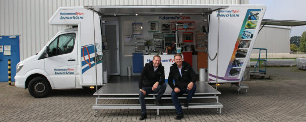 Ein an der Seite geöffneter Lieferwagen mit überdachtem Vordeck, wo an der Stufe zum Deck zwei Männer sitzen und lächeln.