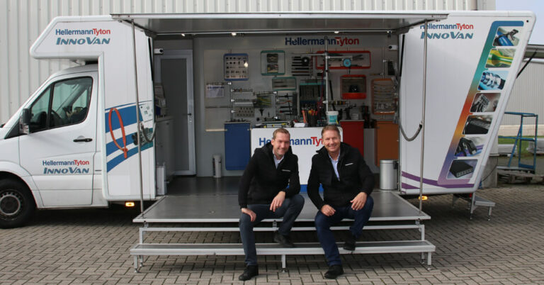 Ein an der Seite geöffneter Lieferwagen mit überdachtem Vordeck, wo an der Stufe zum Deck zwei Männer sitzen und lächeln.