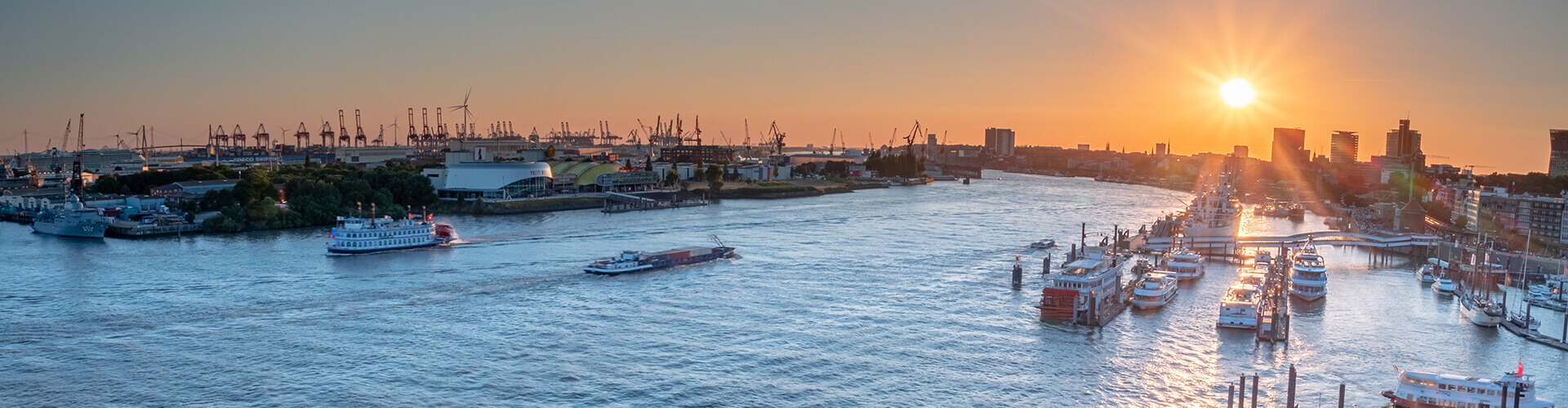 Die Elbe in Hamburg bei Sonnenuntergang mit vorbeifahrenden Schiffen, Containerterminal, zahlreichen Hafenkränen und der Skyline der Hansestadt. Das warme Abendlicht spiegelt sich auf dem Wasser des Hamburger Hafens