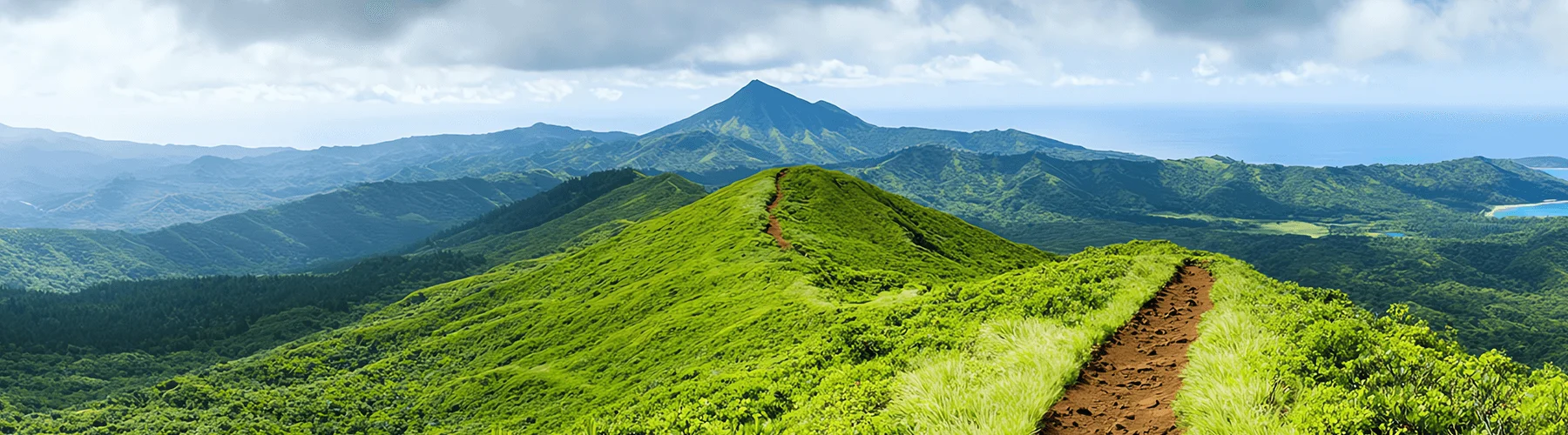 Eine begrünte Berglandschaft mit einem Wanderpfad mittig auf einem Hügel, im Hintergrund am rechten Bildrand ist das Meer.
